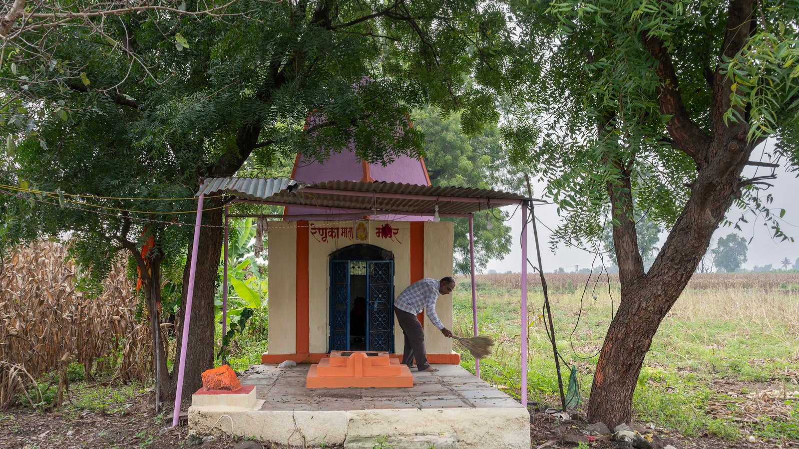 A person is sweeping in front of a small temple surrounded by trees and an open field.