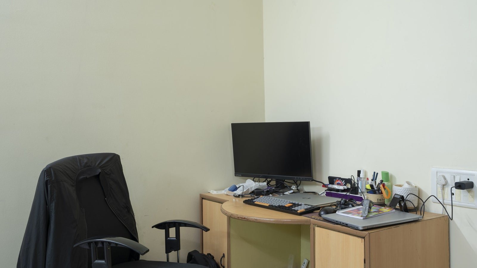 A corner workspace featuring an office chair, a small computer desk with a monitor, keyboard, and various stationery items.
