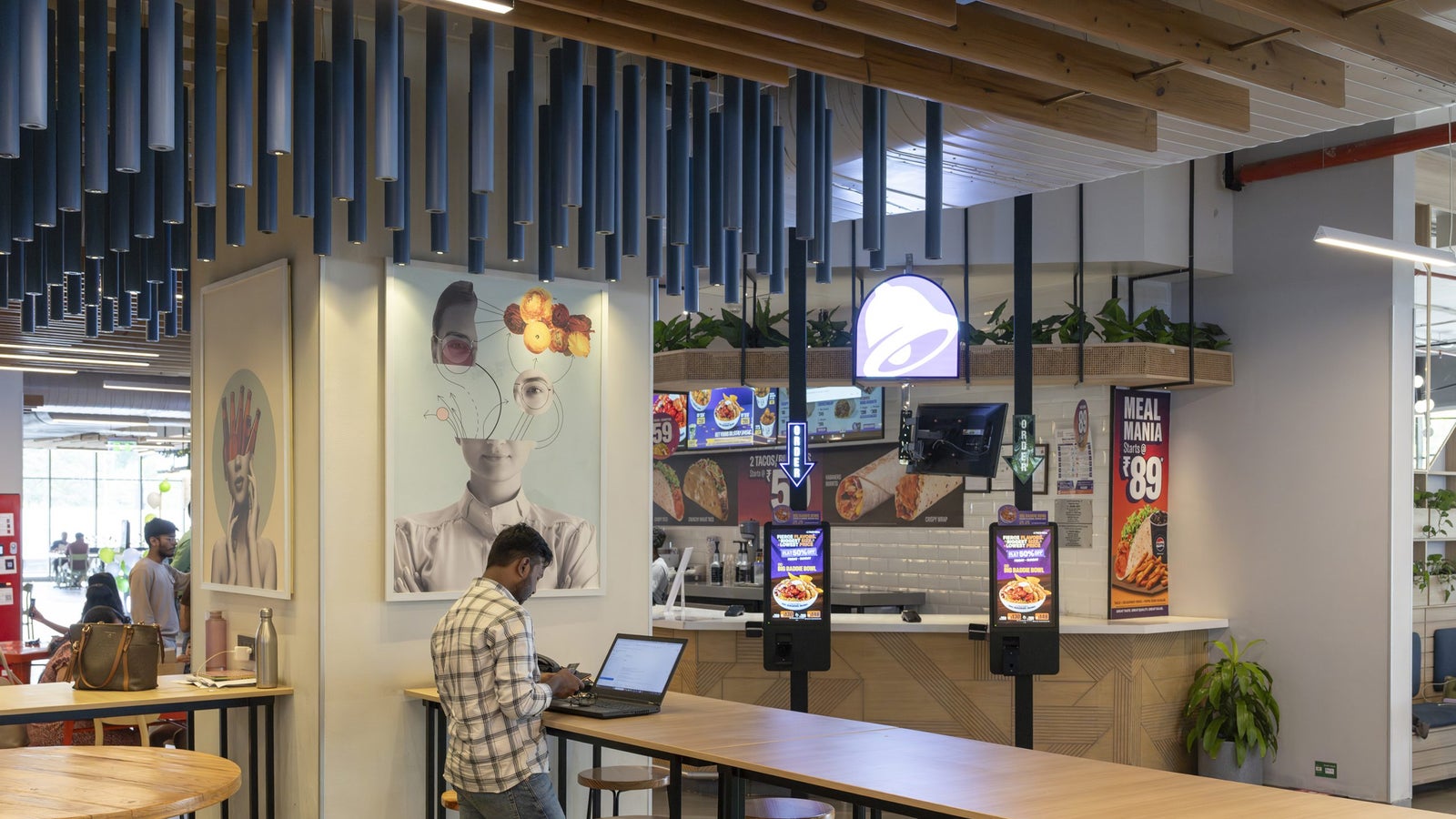 A modern fast-food restaurant interior featuring a digital ordering kiosk, a man using a laptop, and vibrant wall art.
