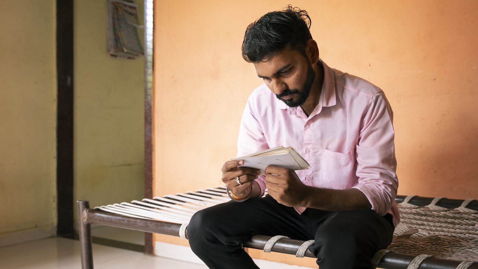 A man in a pink shirt sits on a woven bench, intently reading a notebook in a room with orange walls.