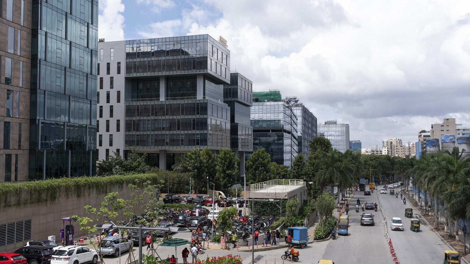 A modern urban street scene featuring buildings, greenery, parked vehicles, and people walking, under a partly cloudy sky.