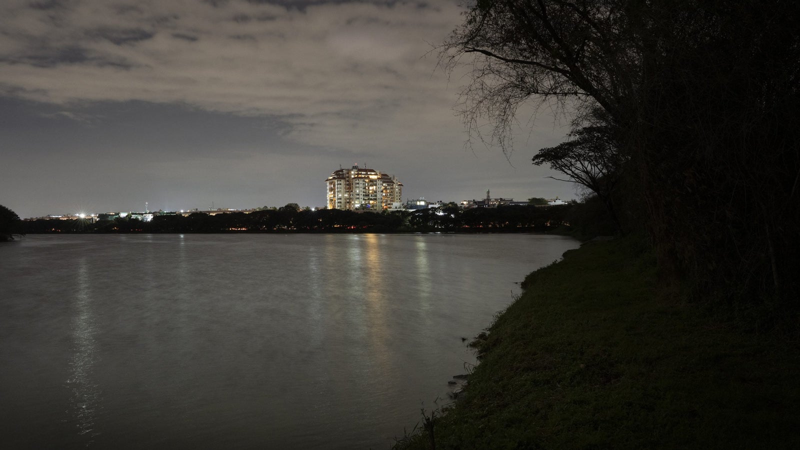 A tranquil river scene at night, with a city skyline illuminated in the background and trees lining the shore.