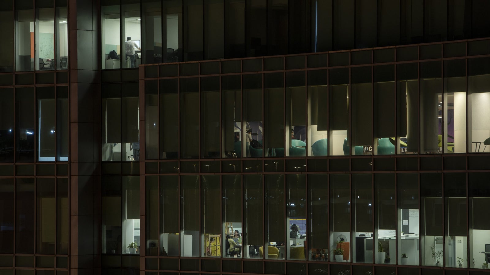 Several office spaces are illuminated at night, showing desks and chairs, with people visible in some of the rooms.