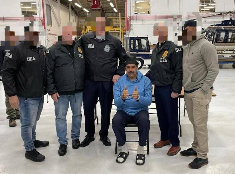 Nicolás Maduro, handcuffed and wearing prison clothes, with Drug Enforcement Agency officers with theiur faces obscured.