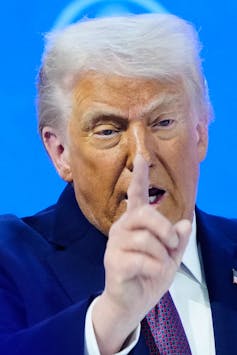 An older man with orange-hued skin and fluffy white hair raises a hand several shades lighter than his face.