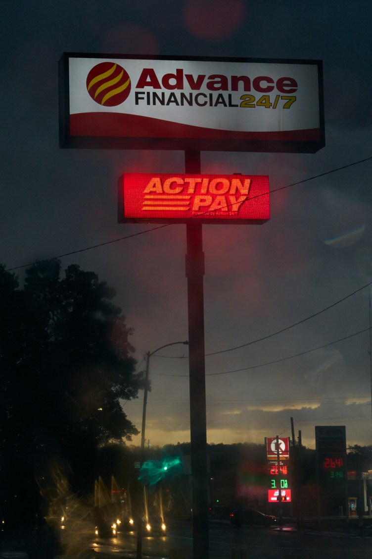 Two large signs on a tall pole rise above stores and streets. The top sign reads “Advance Financial 24/7,” and just below it the second sign reads “Action Pay.”