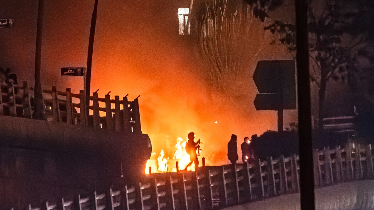 Silhouettes of people near a fire on a bridge, with smoke and orange flames illuminating the scene at night.