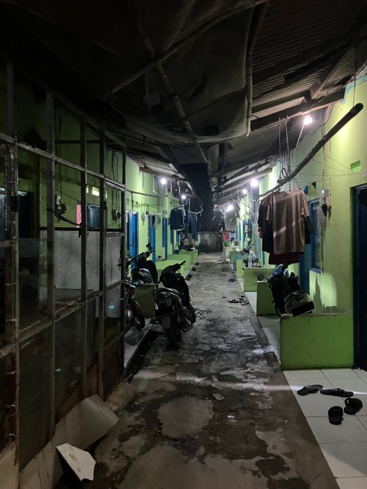 A covered alleyway with a peeling floor, green walls and a sagging ceiling. Clothing hangs from horizontal poles, flip-flops are scattered on the ground and parked motorbikes sit against the walls.