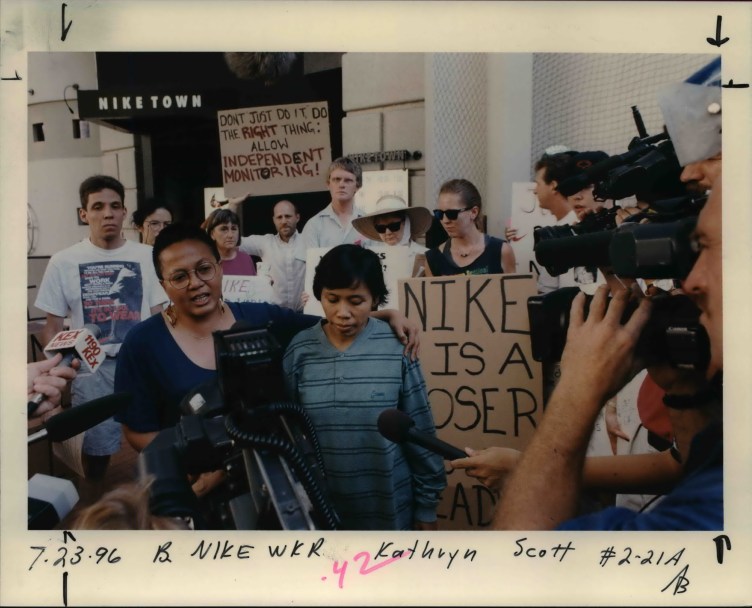 A woman wraps her arm around Cicih’s shoulder in front of a building with a “Nike Town” sign. A crowd of people stand behind the pair holding protest signs, and in front of them are reporters holding out video cameras and microphones. One sign says: “Nike is a loser.” Another reads: “Don’t just do it! Do the right thing: Allow independent monitoring.”