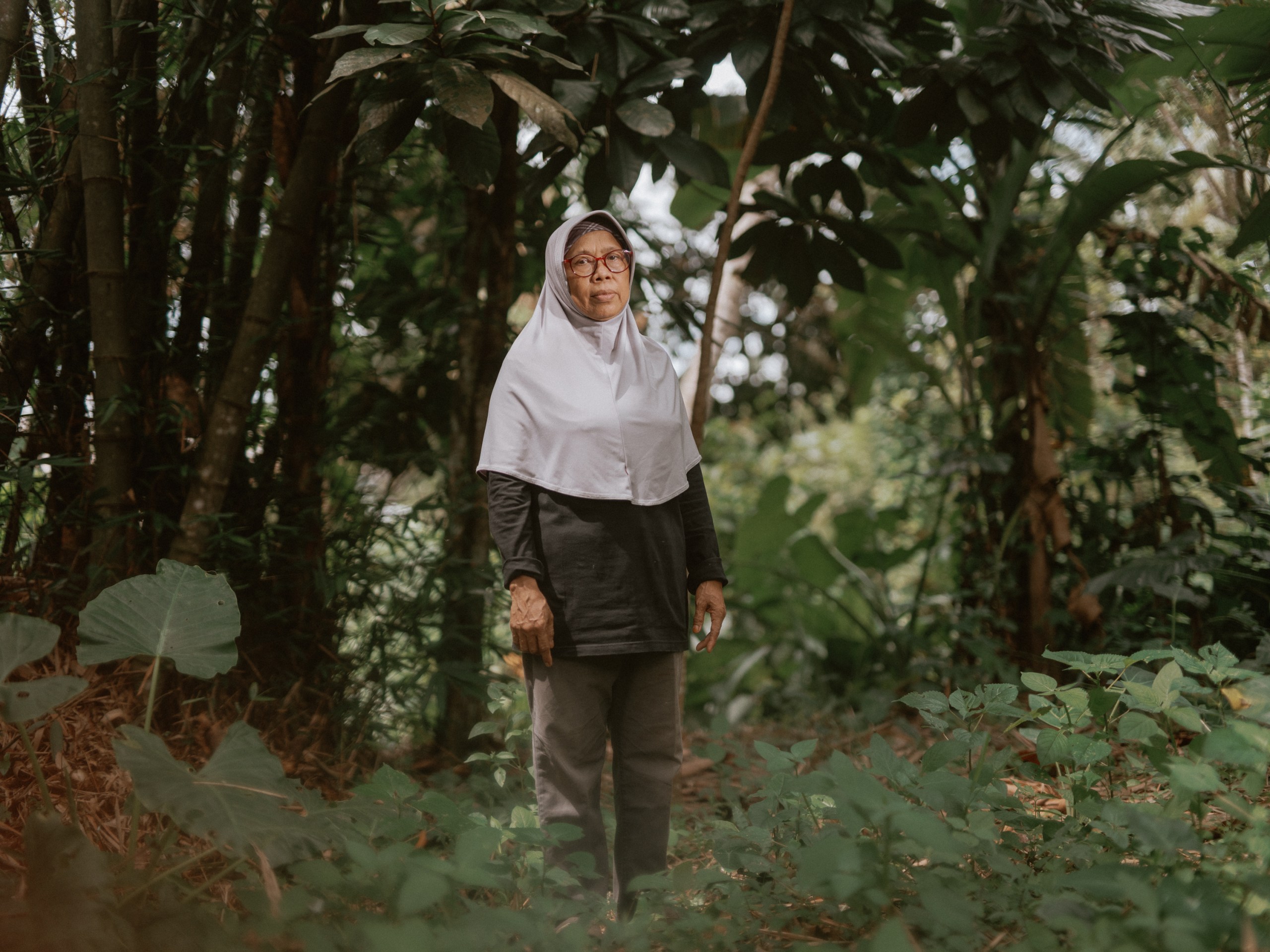 Cicih stands in a forest clearing wearing a light gray hijab and dark gray pants and shirt. Bamboo and other tropical plants grow behind her.