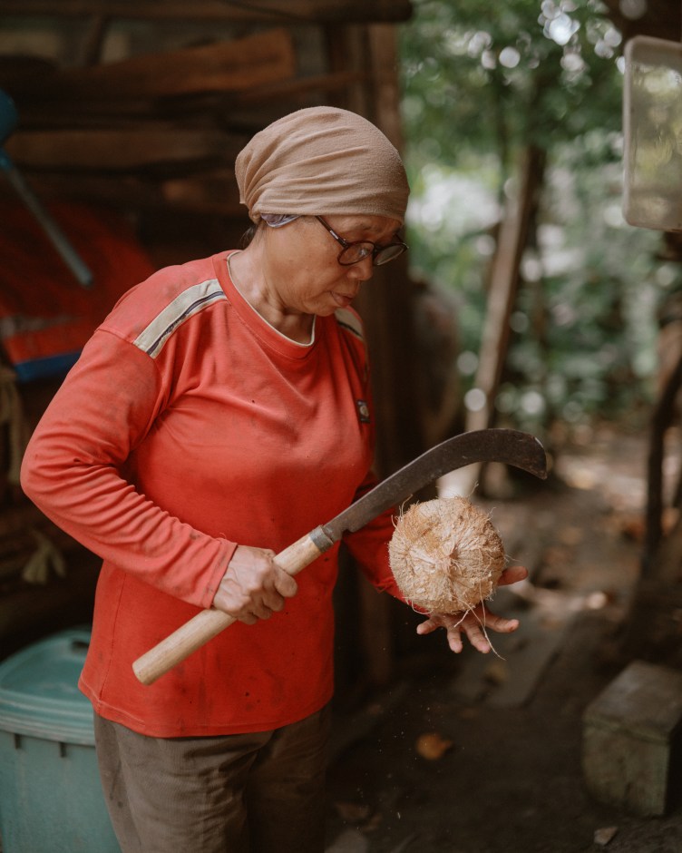Cicih, wearing a beige head wrap and orange long-sleeve shirt, holds a large curved knife in her right hand and a coconut in her left hand.