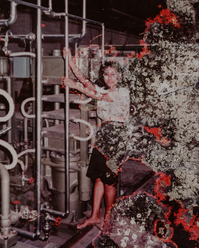 A young Cicih stands holding a pipe attached to factory equipment and leaning back. She has shoulder-length hair, a black skirt, sandals and white shirt. There is red and white damage on the right side of the photograph.