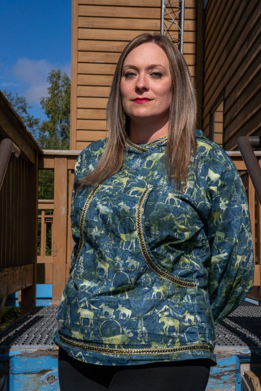 A portrait of a woman standing on an outdoor staircase wearing a jacket with gold accents made out of fabric decorated with a moose and snow motif.