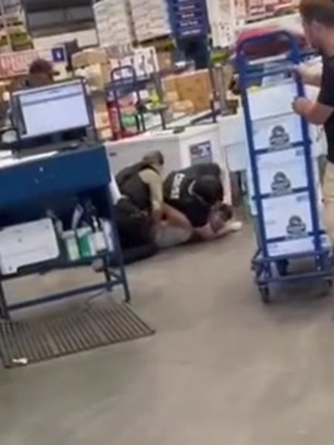 Two men, wearing black armored vests, pin and choke a young man on the ground of a large warehouse store.