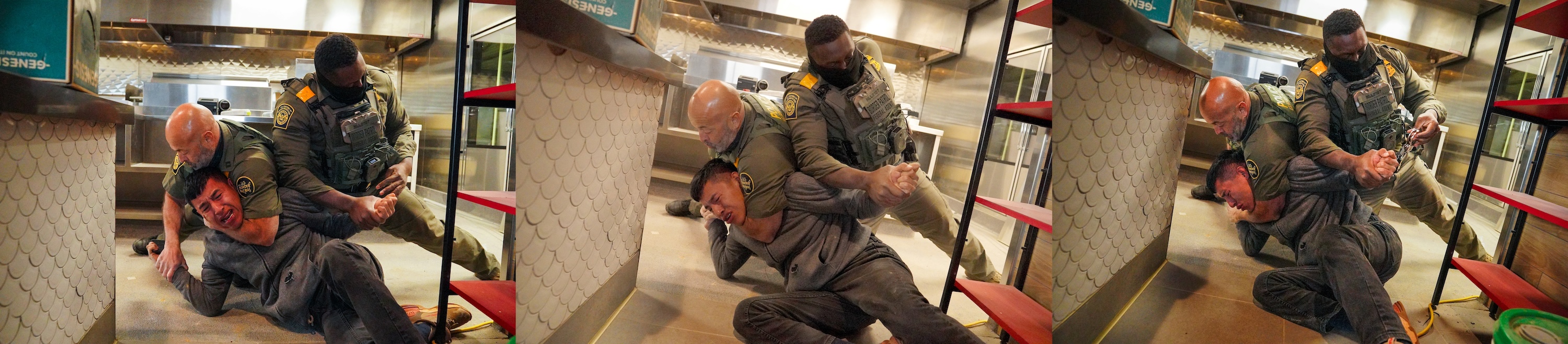 Two uniformed federal officers wearing tactical vests subdue a man wearing a gray sweatshirt and black pants in an industrial kitchen. One officer has his arm around that man’s neck, and the other is holding his wrist.