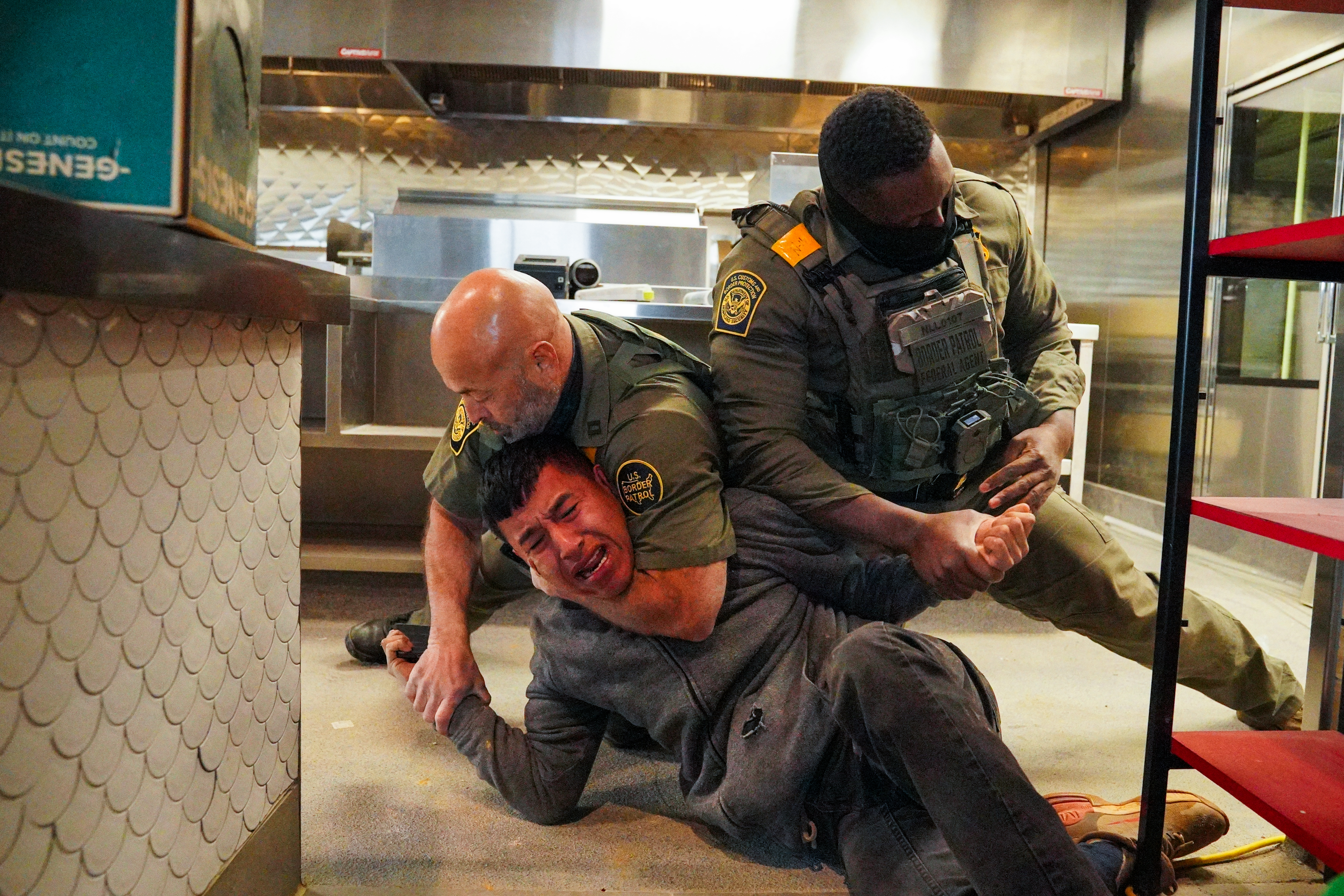 Two uniformed federal officers wearing tactical vests subdue a man wearing a gray sweatshirt and black pants in an industrial kitchen. One officer has his arm around that man’s neck, and the other is holding his wrist.