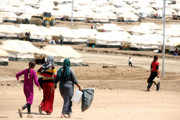Three women walk towards lines  of tents in a refugee camp.