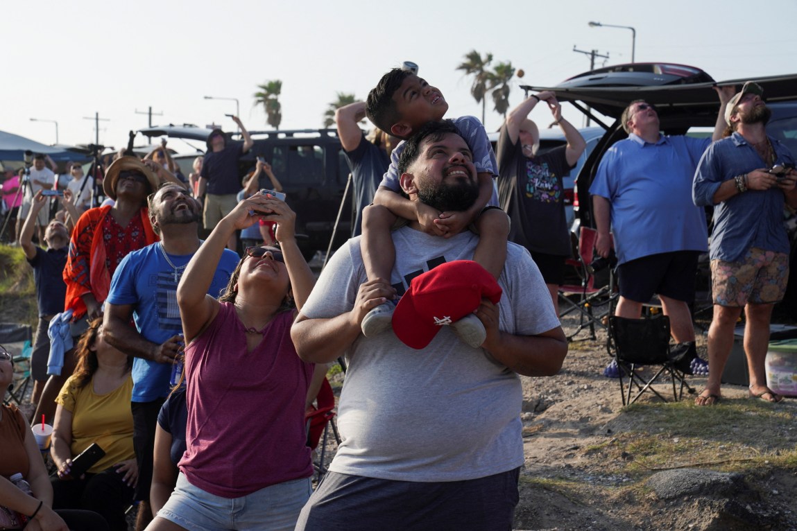A man holds a little boy on his shoulders as they both look up at the sky. Behind them is a big group of people who are also looking up and taking pictures.