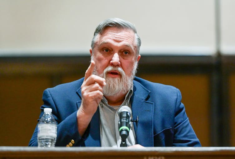 A grey-haired man in a blue suit speaks into a microphone while gesturing with his finger.