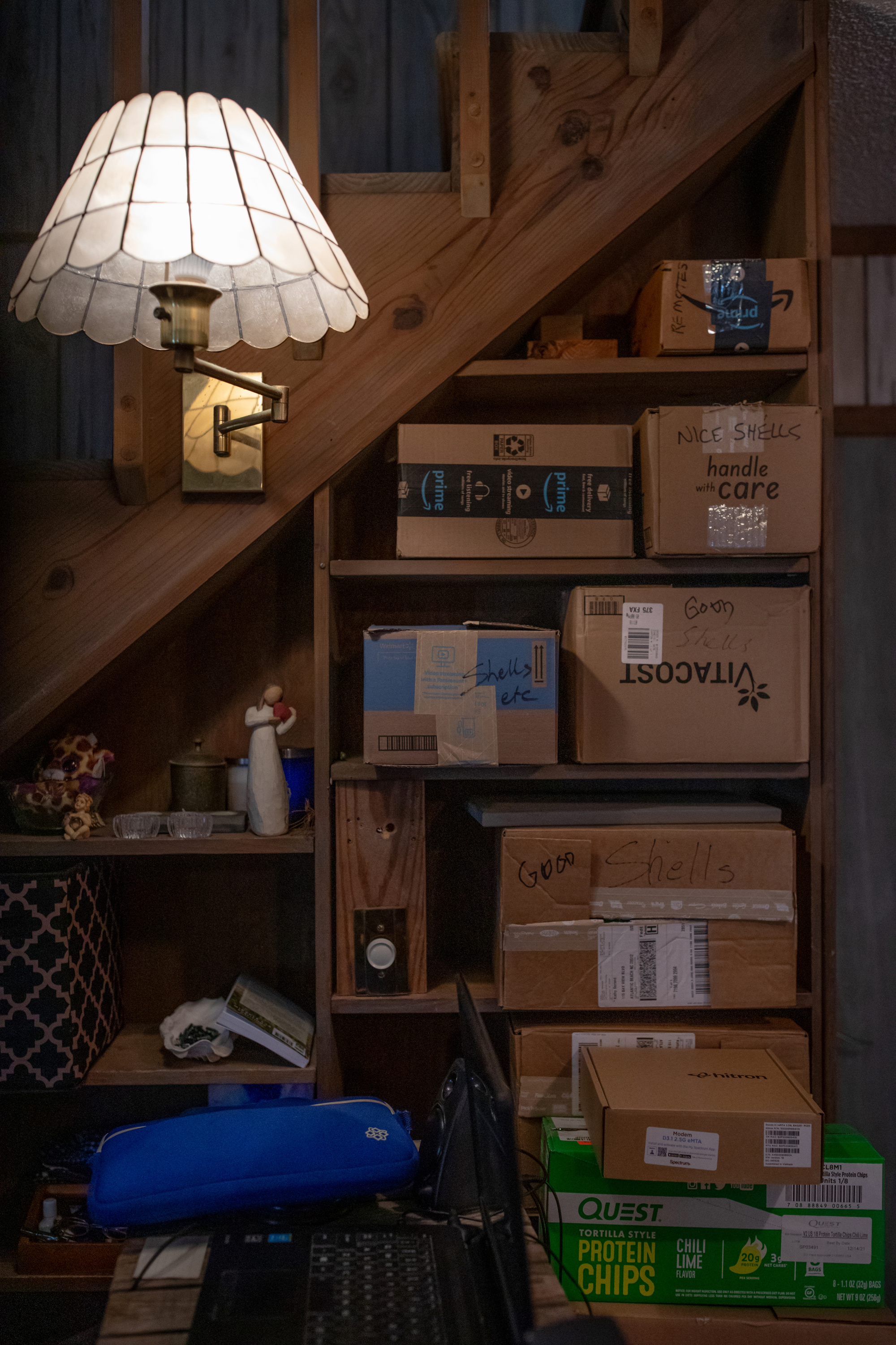 Cardboard boxes fill the space underneath a staircase. Knickknacks sit next to the boxes, and a lit lamp is installed on the staircase.