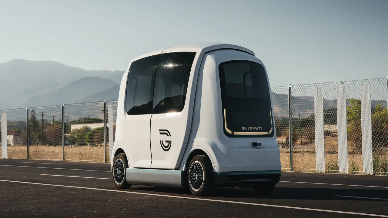 A compact, futuristic autonomous vehicle with a sleek white design, parked on a road with mountains in the background.