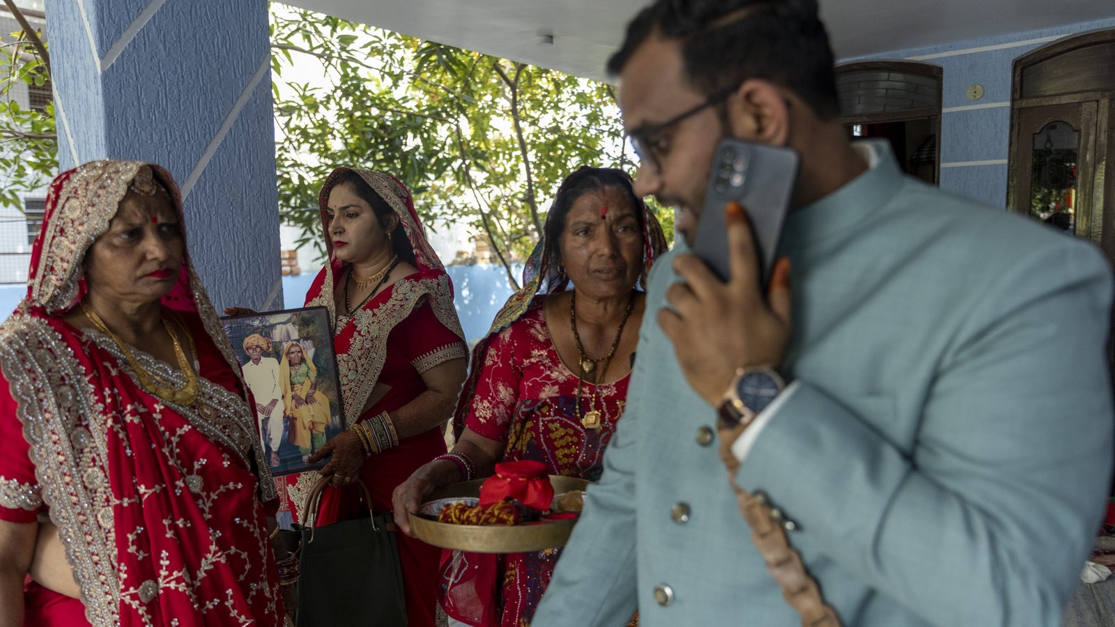 A man in a light blue suit holds a phone, while three women in traditional red attire stand nearby, one holding a photo.