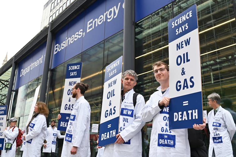 six scientists in white lab coats standing outside building, holding blue white signs highlighting hazard of fossil fuels