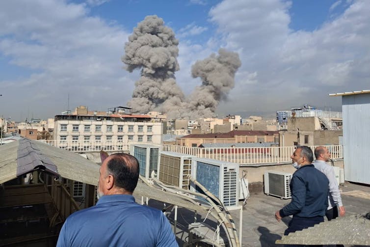 Three iranian men look out from a rooftop as smoke rises from explosions over buildings
