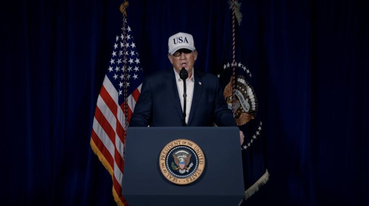 A man in a suit and a baseball cap with USA on it stands at a podium.