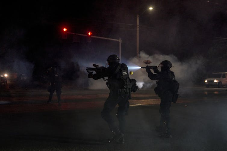 Two police officers dressed in tactical gear aim gun-like weapons into the dark night, with a cloud of tear gas in the background.