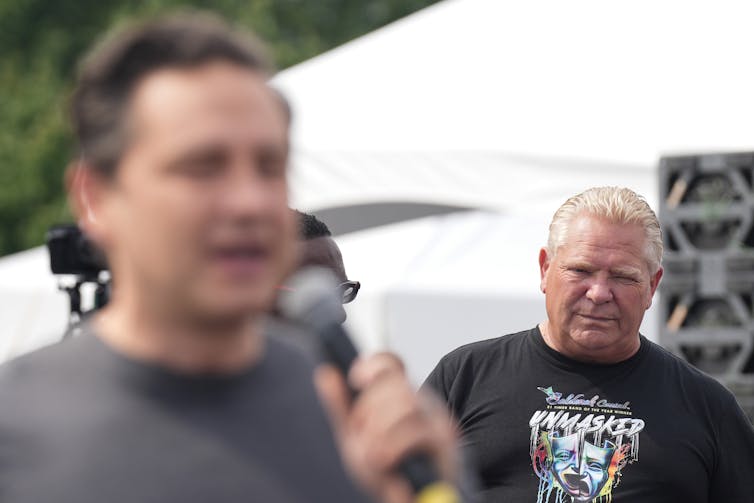 A rotund man with short greyish blond hair watches as a dark-haired man in front of him speaks into a microphone.