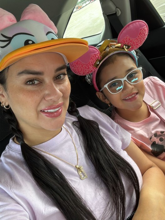 A woman with long black hair and pink lipstick alongside a young girl with white glasses and a pink shirt. Both of them are wearing Disney character hats.