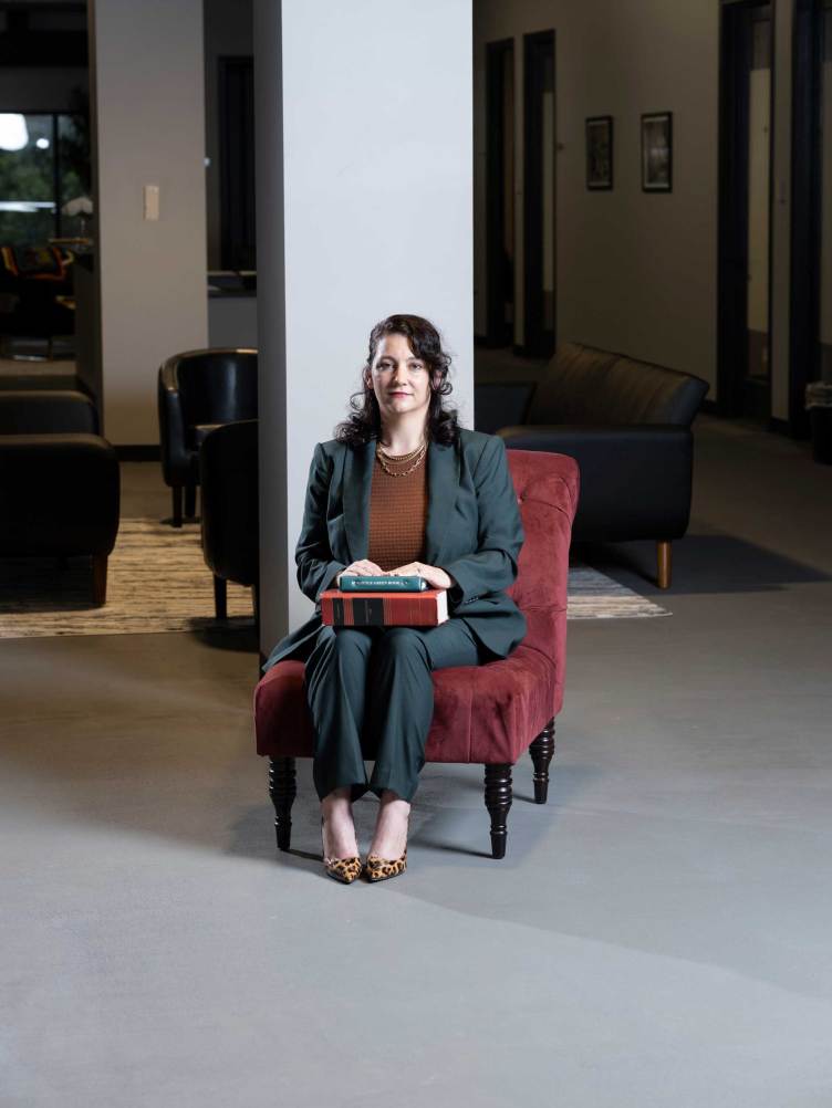 A woman wearing a pantsuit sits on a red velvet chair with two books perched on her lap.