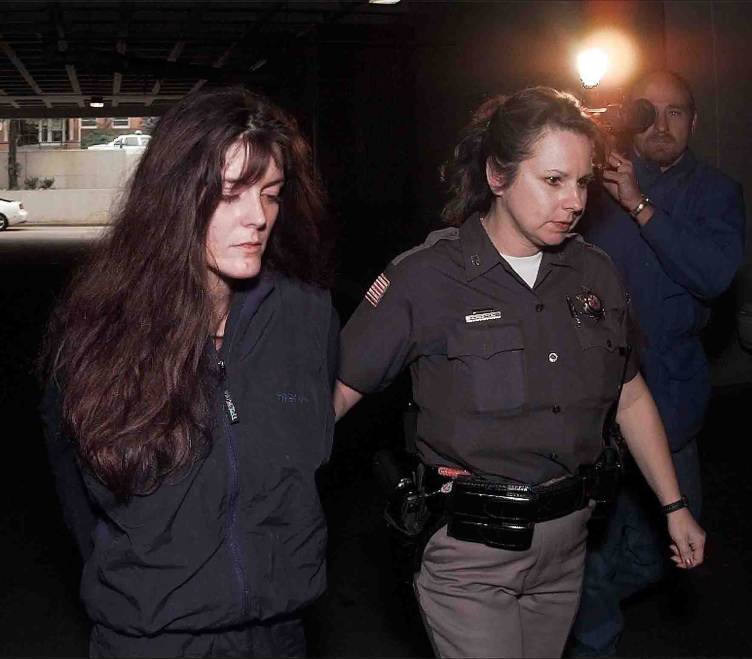 A woman with long brown hair is escorted by a female police officer with a videographer recording their movement in the background.