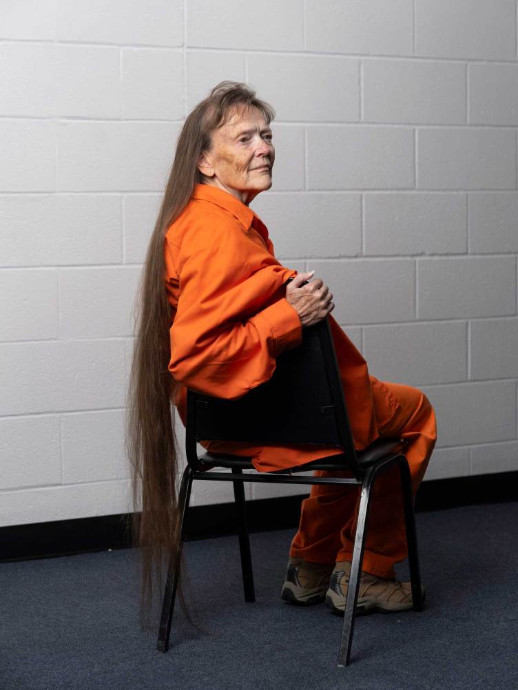 A woman with very long brown hair reaching the ground wearing an all-orange outfit sits on a black metal chair in front of a white, painted cinder block wall.
