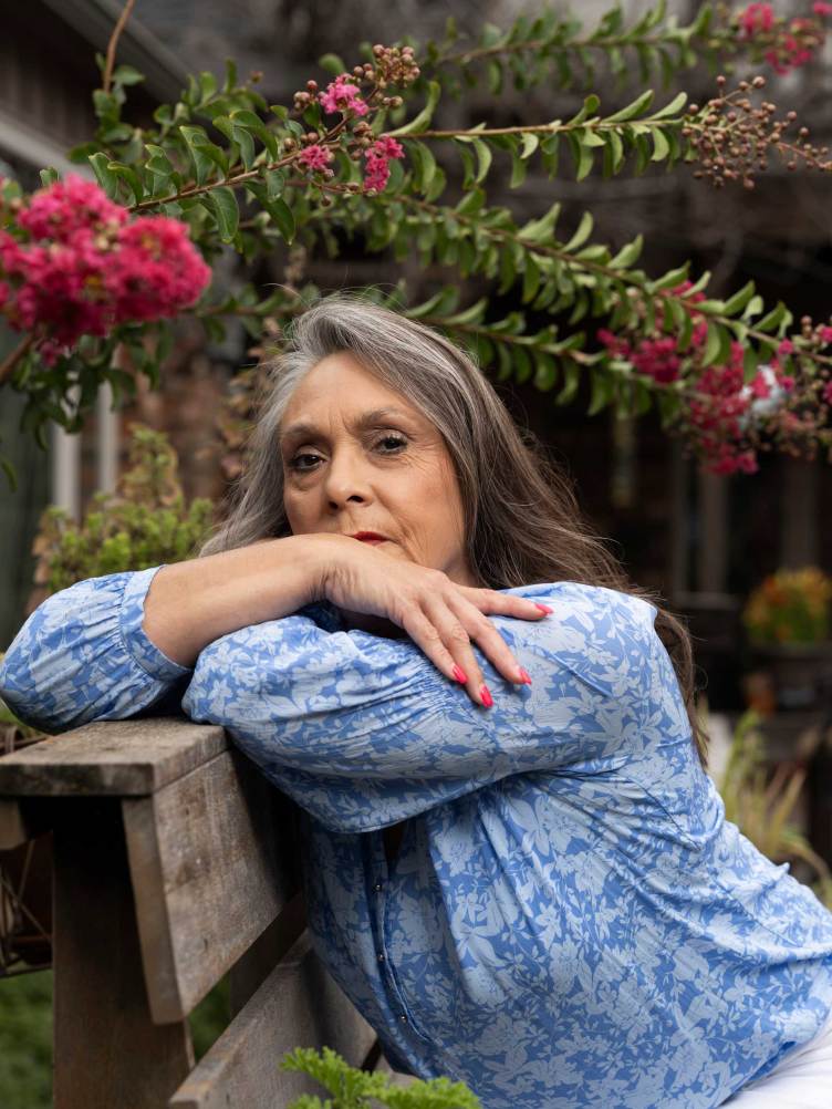 A woman wearing a blue blouse sits on a wooden bench encircled by foliage and pink flowers.