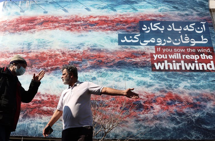 Two men talk in front of a poster reading: If you sow the wind, you will reap the whirlwind.