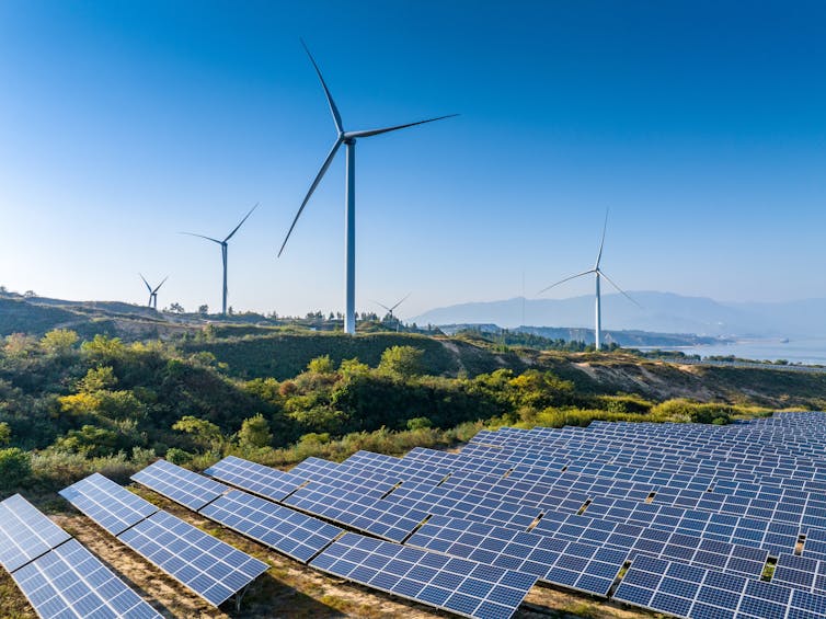 wind and solar farms side by side against a blue sky and mountains in the distance.