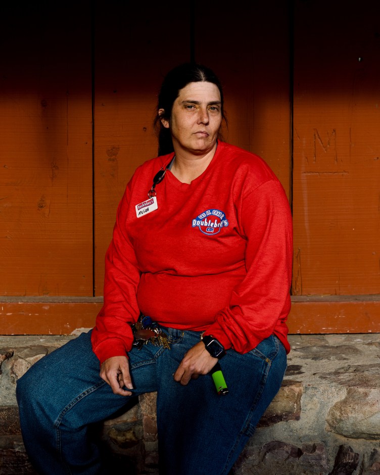 A woman with long black hair, wearing a red long-sleeve shirt and blue jeans, seated on a rock wall against a red, wooden structure.
