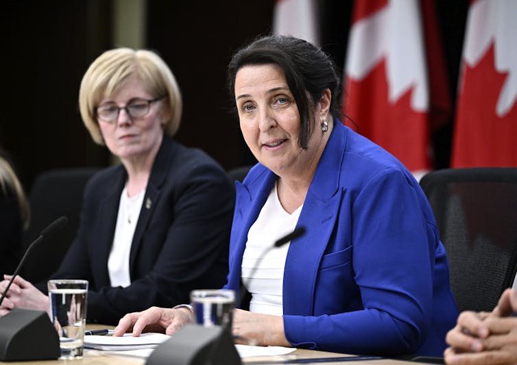 Two women sitting at a conference table and speaking with Canadian flags behind them.