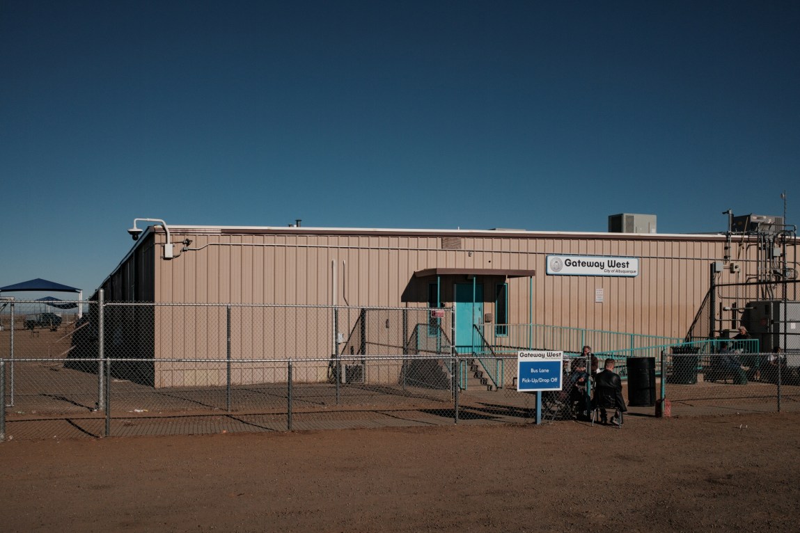 A few people sit outside a large tan building with a turquoise door, surrounded by a chain-link fence.