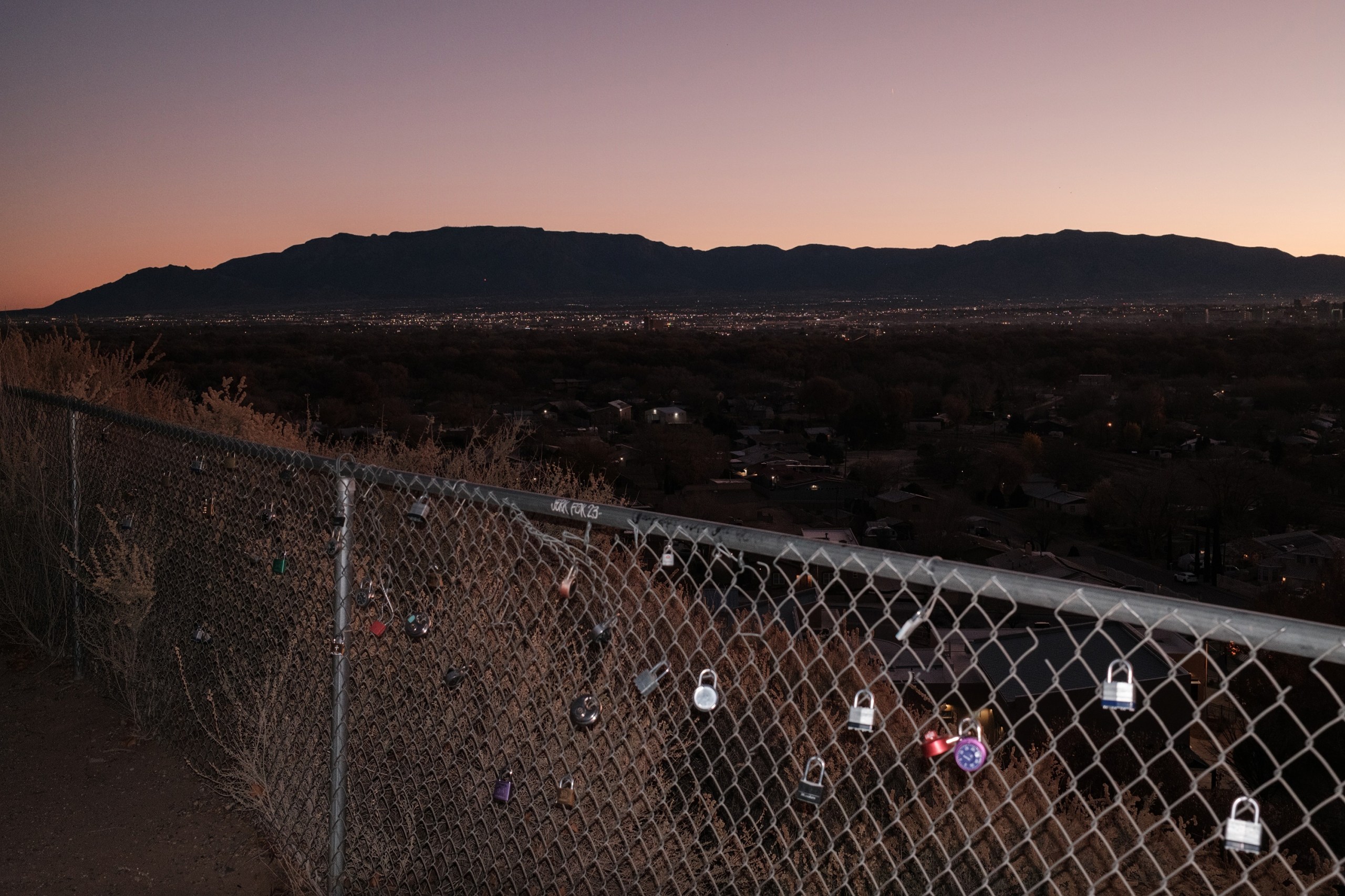 Padlocks clipped to a chain-link fence, with the city of Albuquerque in the distance against a silhouetted ridgeline at dusk.