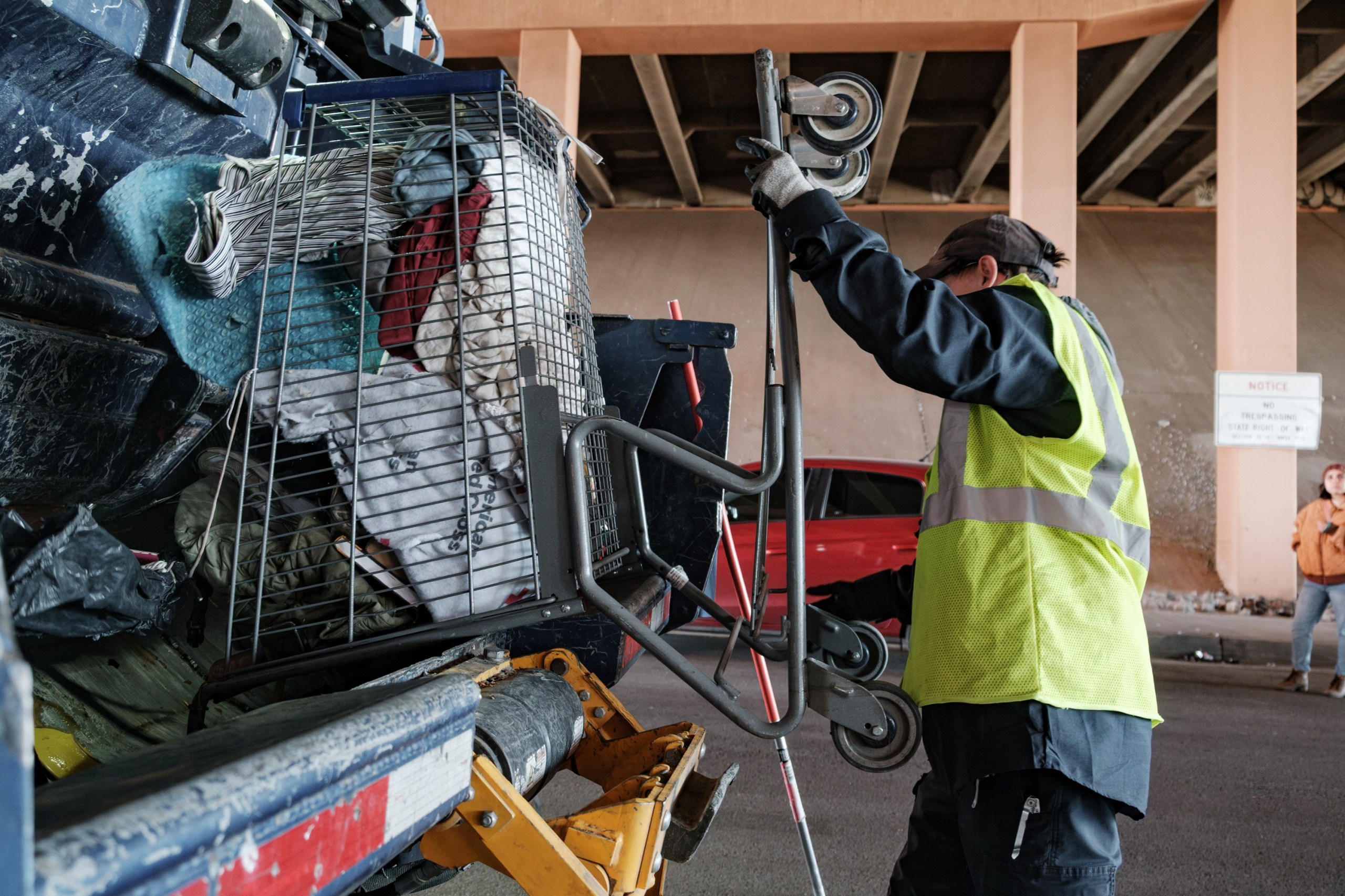 A man wearing a bright yellow vest empties a shopping cart filled with belongings into the compactor of a dump truck under an overpass.