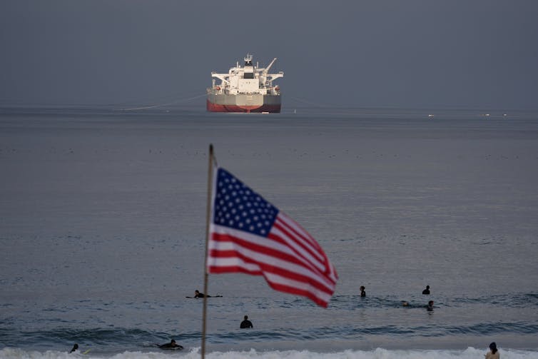 An American flag flies in the foreground while a crude oil tanker sails in the background