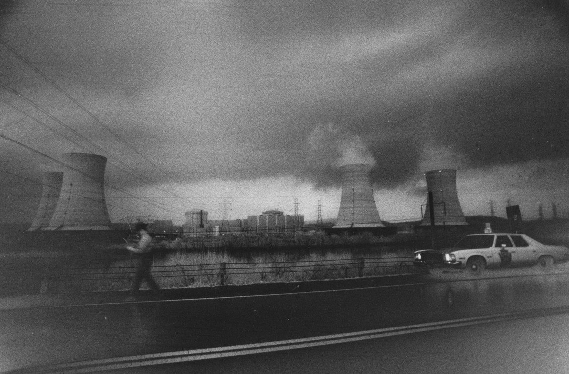 A road with a parked police car and a blurred officer. In the distance are four nuclear cooling towers billowing smoke.
