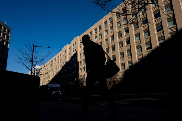 A silhouette of a person wearing a shoulder bag is surrounded by shadow. Behind the person is a large building full of windows and a blue sky.