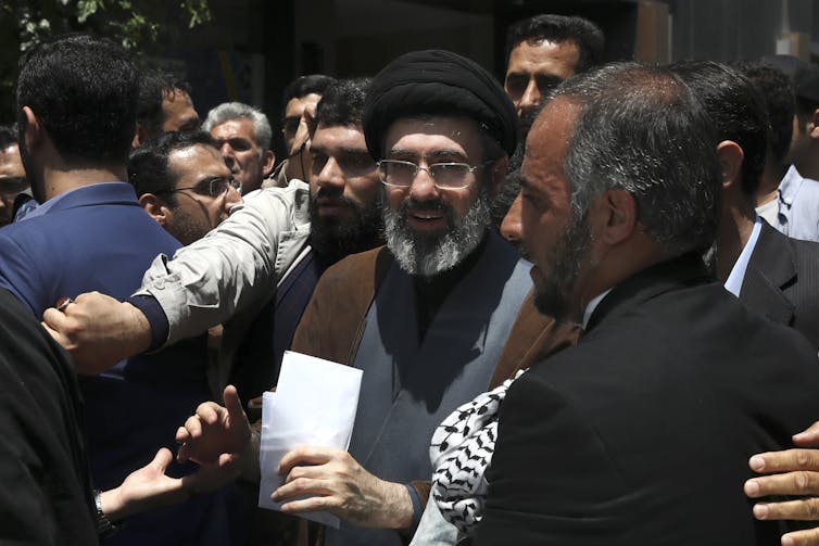 Several men surround a bearded, spectacled man wearing a black headdress.