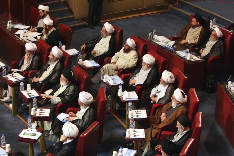 A group of clerics wearing traditional head coverings and long beards sit on seats arranged in concentric rows.