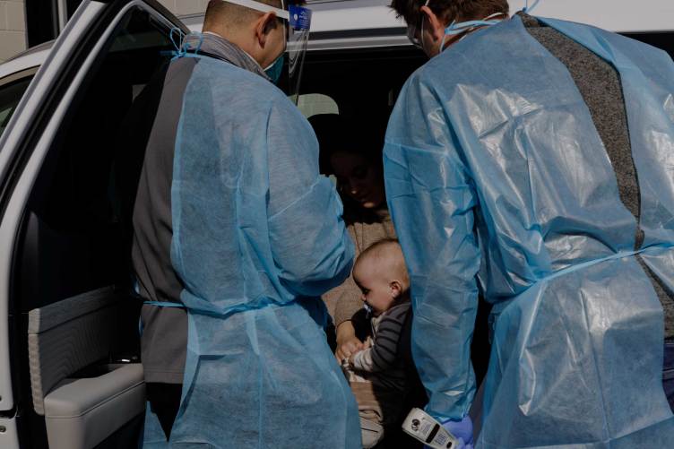 Two physicians wearing gowns, masks and other protective gear stand beside an infant seated in a car with the door open.