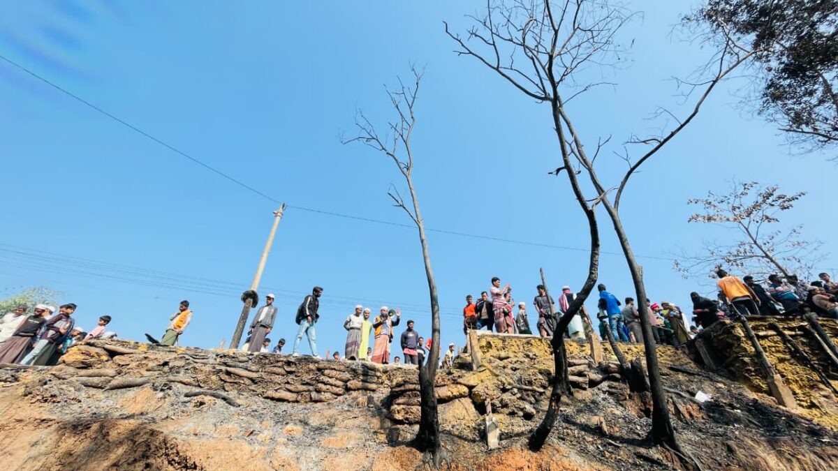 A group of residents stand on a raised embankment looking down at charred ground and burnt trees after a fire destroyed and damaged multiple shelters in Camp 11 in the early morning of 13 February 2026.